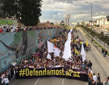 viele Menschen laufen protestierend auf der Straße in Kolumbien und halten ein Banner hoch mit dem Titel #Defendamoslapaz