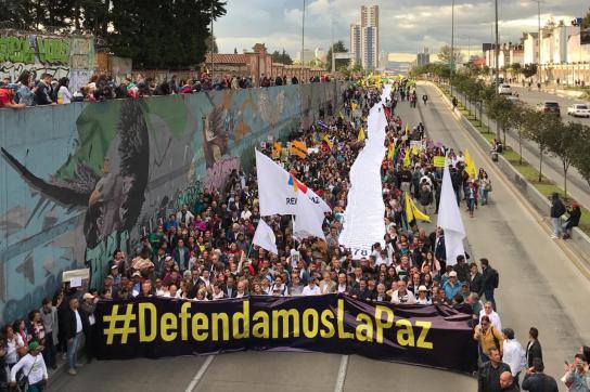 viele Menschen laufen protestierend auf der Straße in Kolumbien und halten ein Banner hoch mit dem Titel #Defendamoslapaz
